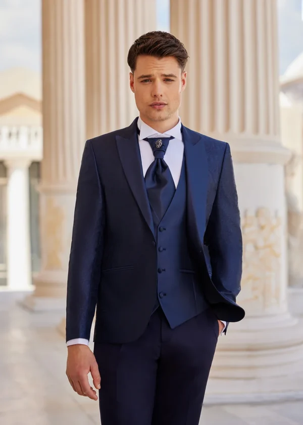 Homme vêtu d’un costume bleu marine raffiné avec des motifs discrets, accompagné d’un gilet et d’une lavallière bleu nuit ornée d’une broche élégante, dans un cadre architectural classique.