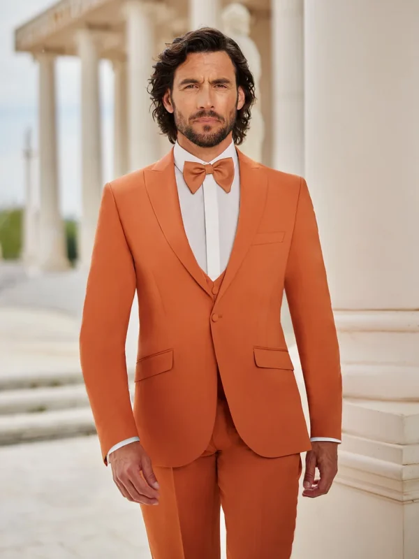 Homme portant un costume couleur terracotta clair, complété par un gilet et un nœud papillon assortis, devant un cadre architectural classique avec colonnes.