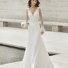 Une mariée portant une robe blanche avec un corsage en dentelle transparente et des manches longues à pois, posant dans un cadre minimaliste en pierre près d’un bassin.
