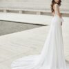 Une mariée de dos en robe blanche avec un dos plongeant en dentelle et une longue traîne fluide posant dans un cadre en pierre minimaliste près de l'eau.