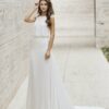 Une mariée portant une robe blanche minimaliste avec un haut fluide sans manches et une jupe droite, posant dans un couloir moderne en pierre.