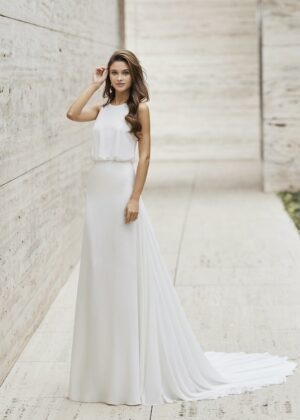 Une mariée portant une robe blanche minimaliste avec un haut fluide sans manches et une jupe droite, posant dans un couloir moderne en pierre.