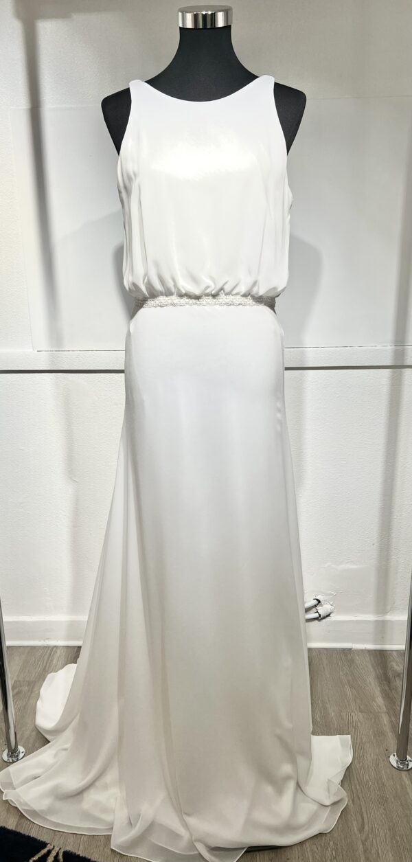 Une robe de mariée blanche minimaliste, avec un haut fluide sans manches et une jupe droite. Elle se distingue par sa simplicité élégante, mise en valeur dans un couloir moderne en pierre.