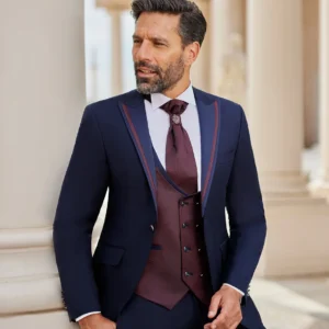 Homme portant un costume bleu marine élégant avec des détails bordeaux, complété par un gilet et une lavallière assortis, posant devant un décor architectural classique.