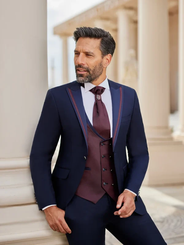 Homme portant un costume bleu marine élégant avec des détails bordeaux, complété par un gilet et une lavallière assortis, posant devant un décor architectural classique.