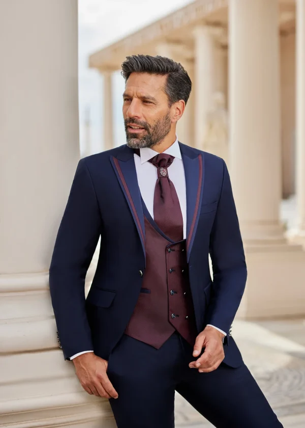 Homme portant un costume bleu marine élégant avec des détails bordeaux, complété par un gilet et une lavallière assortis, posant devant un décor architectural classique.