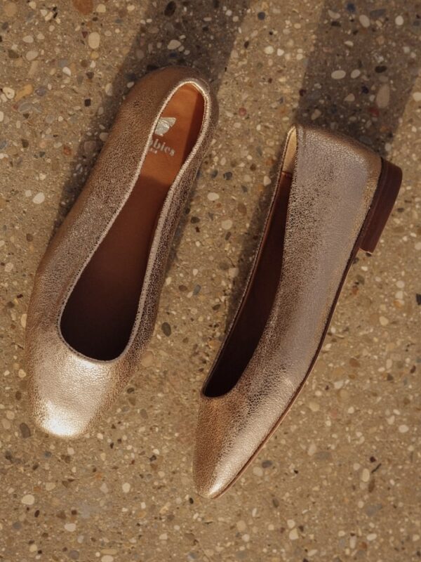 Deux ballerines dorées posées sur un sol en pierre, une vue de dessus dévoilant leur silhouette simple et élégante.