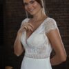 Portrait en plan rapproché d'une femme souriante portant une robe de mariée en dentelle. La photo met en valeur les détails délicats des manches et du décolleté plongeant de la robe, ainsi que l'élégance naturelle de la mariée