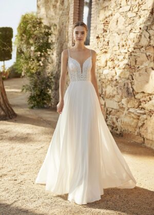 Robe de mariée à bretelles fines avec décolleté en V profond, ornée de dentelle florale délicate, portée par une femme élégante devant un mur en pierre naturel