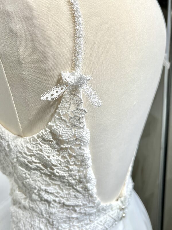 Robe de mariée à bretelles fines avec décolleté en V profond, ornée de dentelle florale délicate, portée par une femme élégante devant un mur en pierre naturel