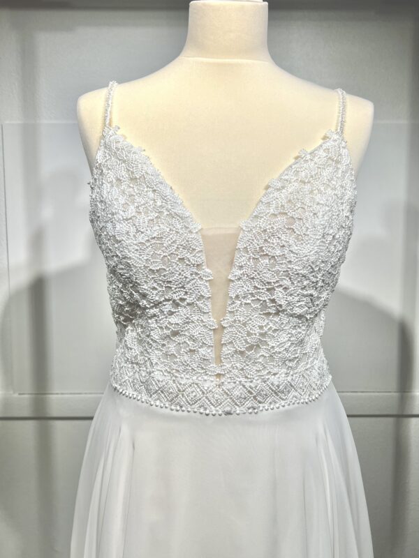 Robe de mariée à bretelles fines avec décolleté en V profond, ornée de dentelle florale délicate, portée par une femme élégante devant un mur en pierre naturel