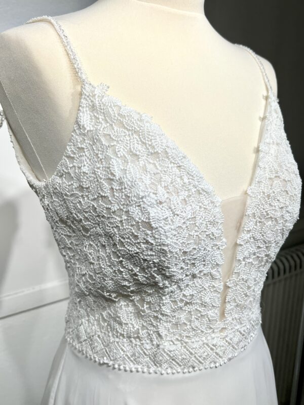 Robe de mariée à bretelles fines avec décolleté en V profond, ornée de dentelle florale délicate, portée par une femme élégante devant un mur en pierre naturel