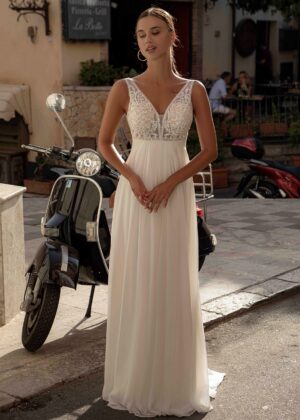 Mariée debout devant un scooter vintage noir, vêtue d'une robe fluide avec un corsage en dentelle et un décolleté en V, dans un cadre de village ensoleillé
