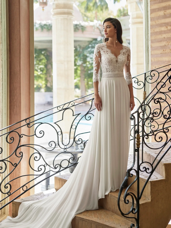 Mariée élégante portant une robe de mariée avec un haut en dentelle à manches longues et une jupe fluide, posant sur un escalier orné de fer forgé