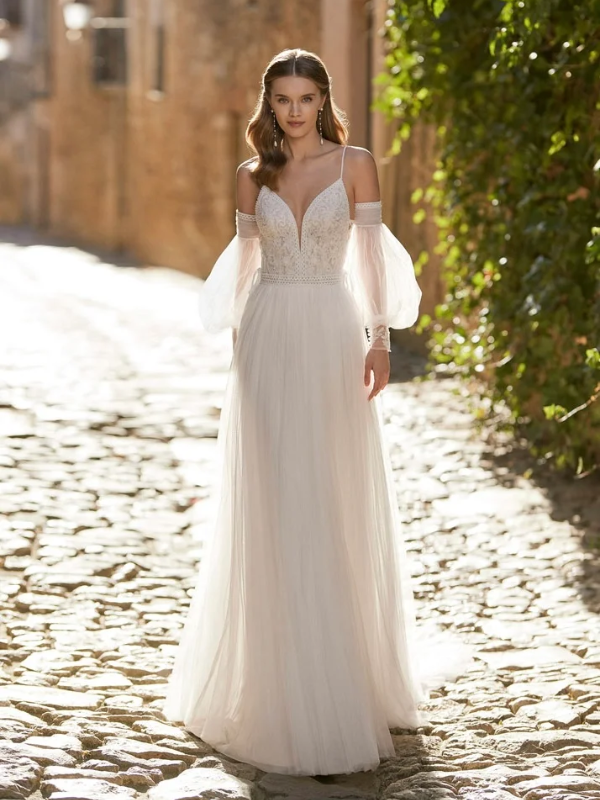 Mariée portant une robe fluide avec un décolleté en V et des manches bouffantes transparentes, marchant dans une rue pavée baignée de lumière naturelle