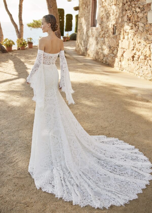 Vue arrière d'une robe de mariée fluide en dentelle avec un dos ouvert, portée par une mariée accompagnée d'un homme en costume beige