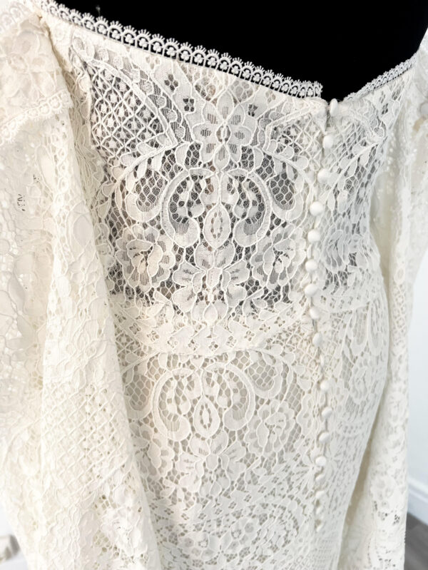 Robe de mariée en dentelle à manches trois-quarts avec détails floraux, avec une mariée élégante en pleine conversation avec un homme en costume beige