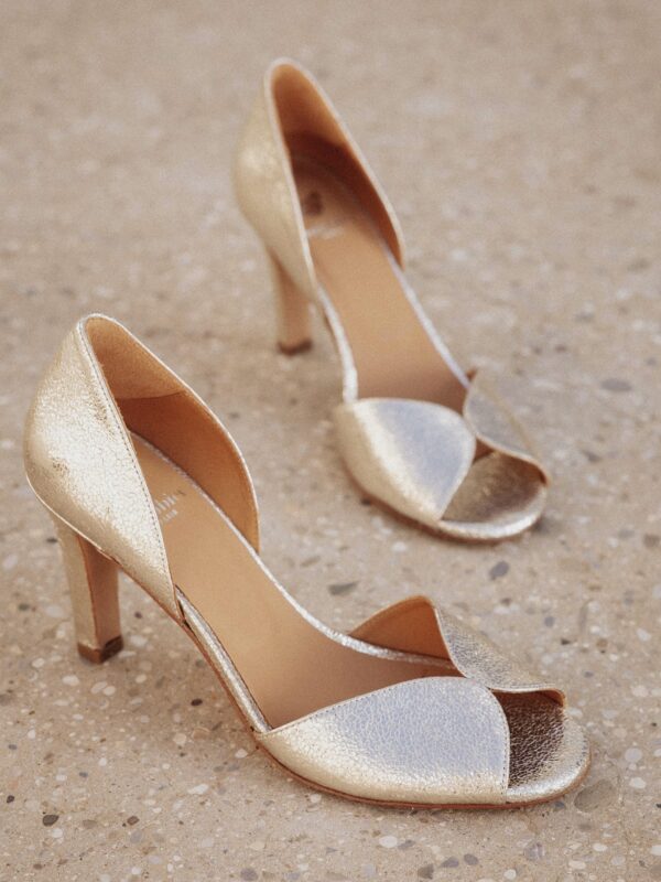 Deux escarpins dorés ouverts posés sur un sol en pierre, avec un design à talons fins et une ouverture élégante sur le devant.