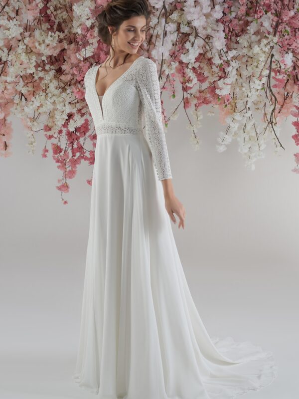 Femme souriante vêtue d’une robe de mariée fluide, avec un corsage en dentelle et manches longues, posant devant une cascade de fleurs roses et blanches suspendues. La robe présente un décolleté en V et une taille marquée par une délicate ceinture de dentelle