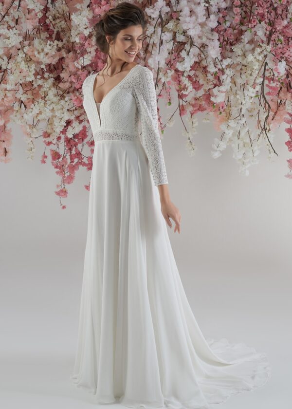 Femme souriante vêtue d’une robe de mariée fluide, avec un corsage en dentelle et manches longues, posant devant une cascade de fleurs roses et blanches suspendues. La robe présente un décolleté en V et une taille marquée par une délicate ceinture de dentelle