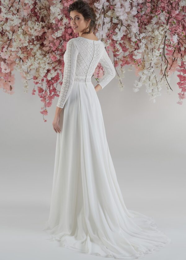 Vue arrière d’une robe de mariée fluide, avec un corsage en dentelle complexe et des manches longues, portée par une femme posant devant une cascade de fleurs suspendues dans des tons roses et blancs. La robe présente un dos légèrement ouvert avec des détails en dentelle et une jupe élégante tombant en douceur