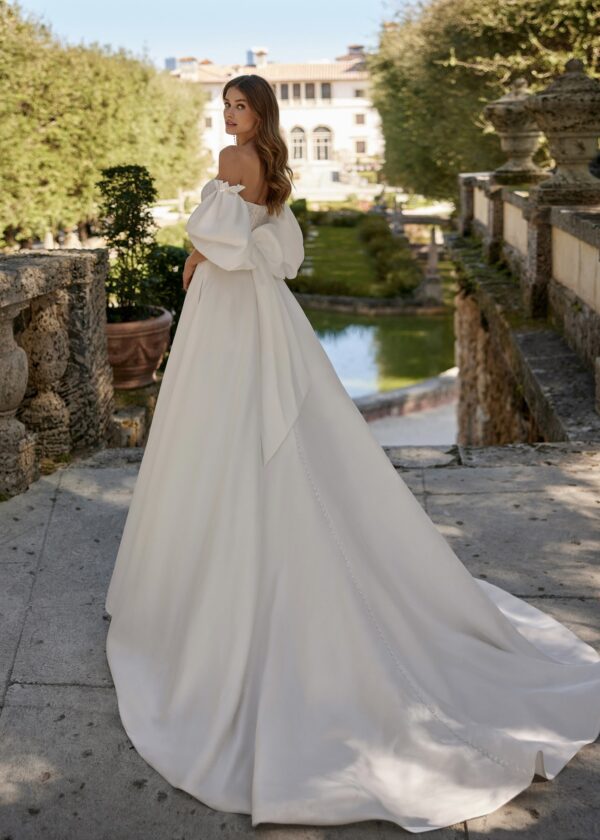 Vue de dos d'une mariée portant une robe volumineuse avec manches bouffantes et un nœud élégant, dans un jardin majestueux