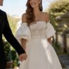 Mariée souriante portant une robe de mariée avec un corsage en dentelle et des manches bouffantes, tenant la main de son partenaire dans un cadre extérieur élégant