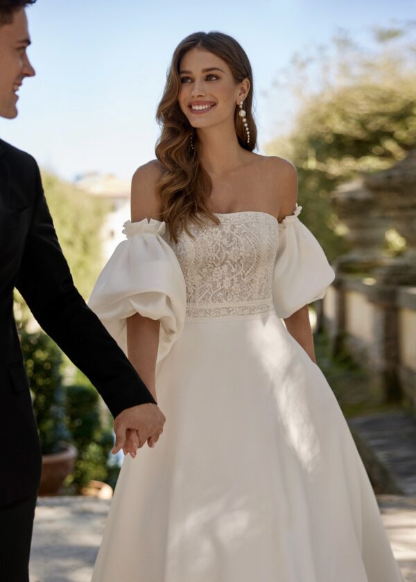 Mariée souriante portant une robe de mariée avec un corsage en dentelle et des manches bouffantes, tenant la main de son partenaire dans un cadre extérieur élégant