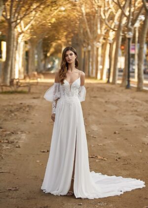 Une femme en robe de mariée bohème blanche avec un décolleté plongeant en V, des manches transparentes en tulle et une jupe fluide fendue, posant sur un chemin bordé d'arbres.