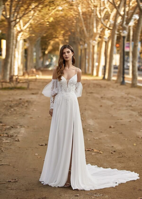 Une femme en robe de mariée bohème blanche avec un décolleté plongeant en V, des manches transparentes en tulle et une jupe fluide fendue, posant sur un chemin bordé d'arbres.