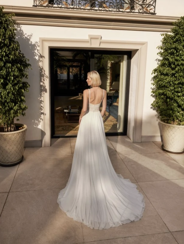 Vue de dos d'une mariée portant une robe fluide avec des bretelles en dentelle et une longue traîne légère, se tenant sur une terrasse élégante devant une porte vitrée.
