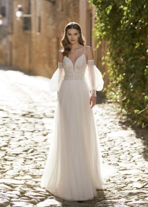 Mariée portant une robe fluide avec un décolleté en V et des manches bouffantes transparentes, marchant dans une rue pavée baignée de lumière naturelle