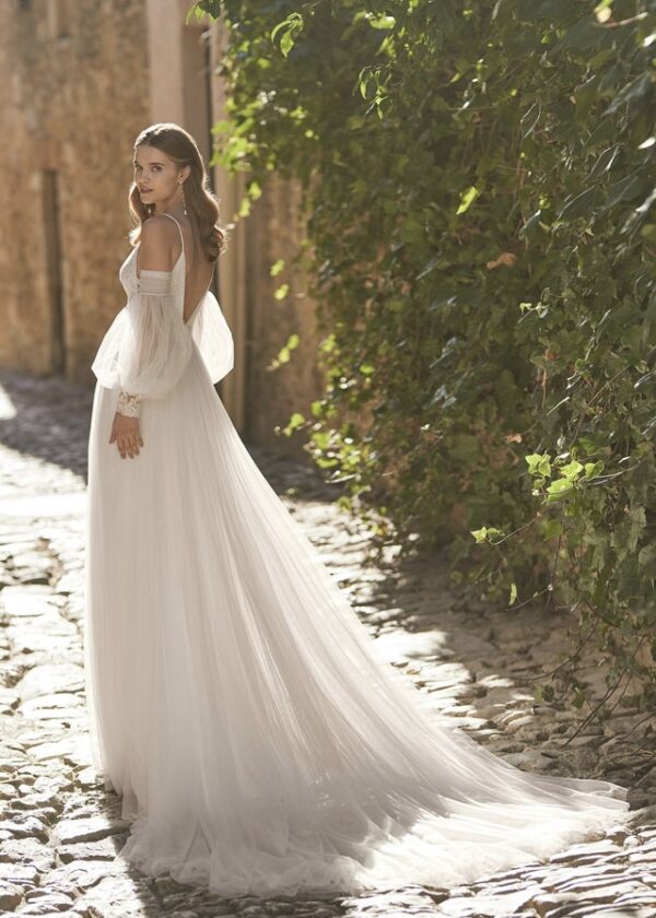 Vue de dos d'une mariée portant une robe fluide avec des manches bouffantes transparentes et une longue traîne, marchant dans une rue pavée bordée de verdure