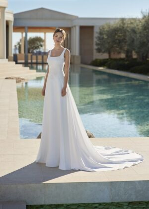 Une mariée portant une robe de mariée blanche avec une coupe droite et un col carré, posant élégamment devant une piscine à l'architecture classique.
