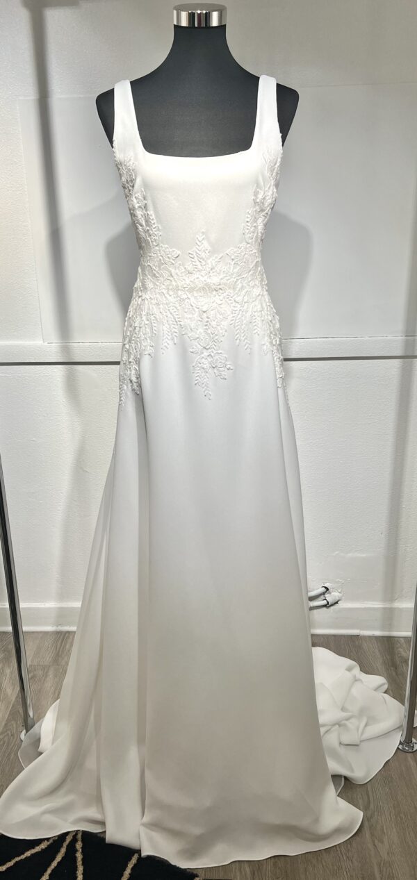 Cette robe de mariée allie minimalisme et élégance avec son corsage ajusté à col carré et sa jupe fluide prolongée d’une traîne légère. Son design épuré s’harmonise parfaitement avec un cadre méditerranéen raffiné, idéal pour une mariée au style intemporel.