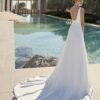 Une mariée de dos en robe de mariée fluide avec un dos nu échancré et une longue traîne, posant près d'une piscine dans un décor méditerranéen.