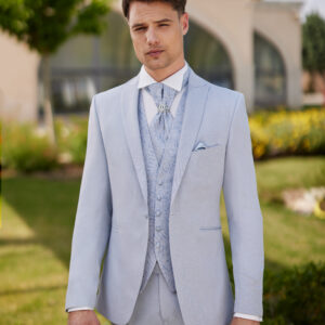 Homme vêtu d’un costume bleu pâle élégant, assorti d’un gilet et d’une lavallière brodée, orné d’une broche, posant devant un décor naturel et architectural.