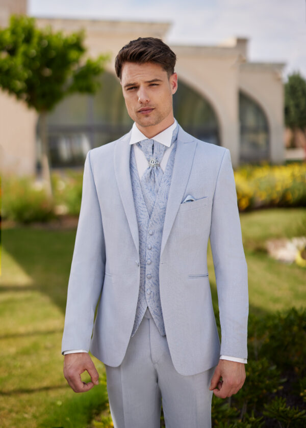 Homme vêtu d’un costume bleu pâle élégant, assorti d’un gilet et d’une lavallière brodée, orné d’une broche, posant devant un décor naturel et architectural.