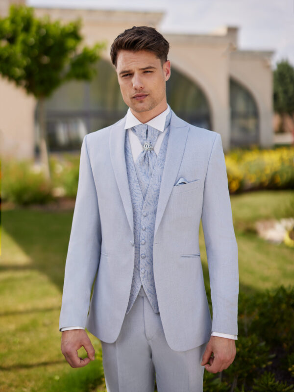 Homme vêtu d’un costume bleu pâle élégant, assorti d’un gilet et d’une lavallière brodée, orné d’une broche, posant devant un décor naturel et architectural.