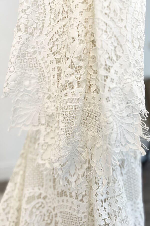 Robe de mariée en dentelle à manches trois-quarts avec détails floraux, avec une mariée élégante en pleine conversation avec un homme en costume beige