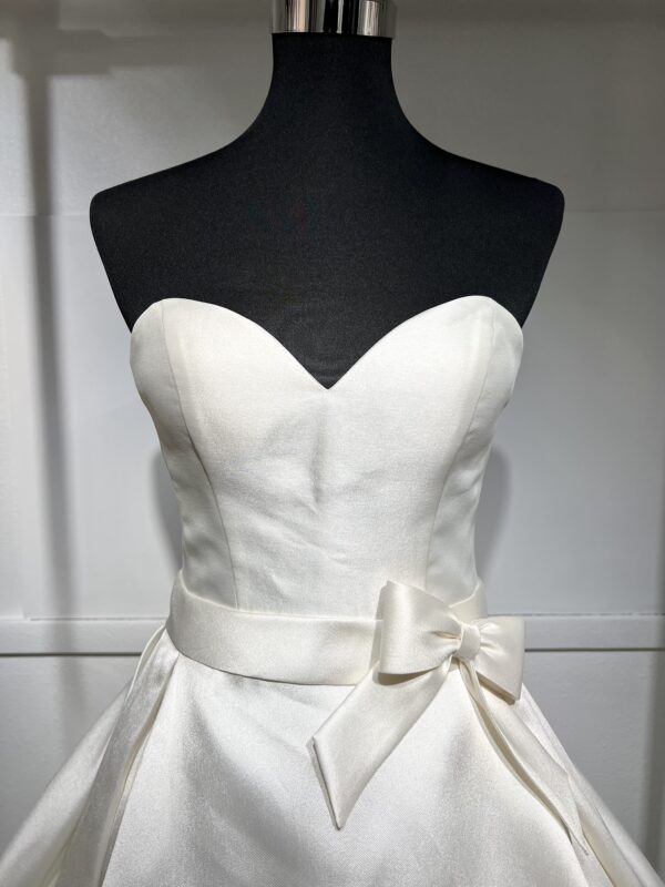 Robe de mariée Divina Sposa coupe princesse en satin blanc, bustier cœur sans bretelles, ceinture à nœud et jupe fluide jusqu’au sol.