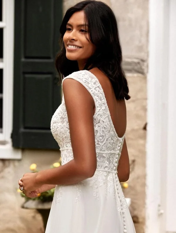 Gros plan de profil d'une mannequin souriante mettant en valeur le bustier en dentelle florale avec mancherons et le décolleté de dos arrondi de la robe de mariée 44275 Justin Alexander.