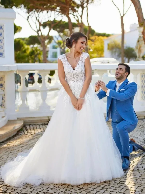 Une mariée de dos en robe blanche avec un dos nu orné de dentelle florale et une longue traîne fluide, posant sur des marches en pierre dans un jardin décoré de statues.