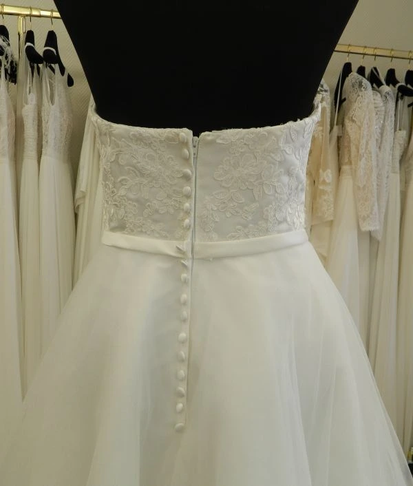 Zoom technique sur le dos du bustier en dentelle et la boutonnière de la robe de mariée courte modèle NC2924 par Nana Couture au showroom.