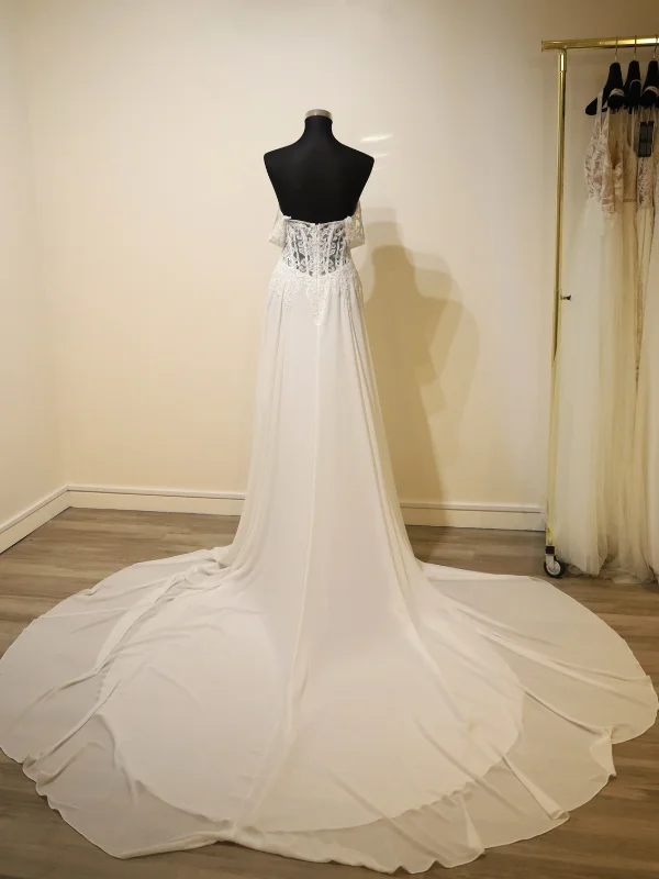 Vue de dos détaillée de la robe de mariée 'Anahi' d'Adriana Alier sur un portant, mettant en valeur le dos en dentelle florale travaillée avec une rangée verticale de boutons nacrés, les petites manches capelines et une longue traîne en mousseline fluide étalée au sol.