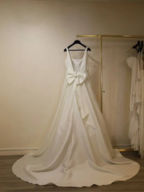 Vue de dos d'une robe de mariée sur cintre, mettant en valeur un élégant nœud décoratif au bas du dos nu et une traîne fluide.