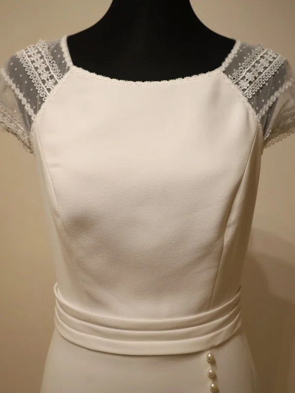 Gros plan sur le bustier d'une robe de mariée, mettant en valeur les manches en dentelle transparente, les détails perlés et la ceinture drapée.