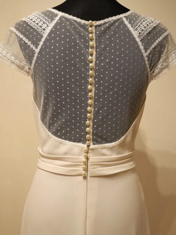 Gros plan sur le dos d'une robe de mariée, mettant en avant un empiècement en dentelle transparente, une rangée verticale de boutons perlés et une ceinture drapée.