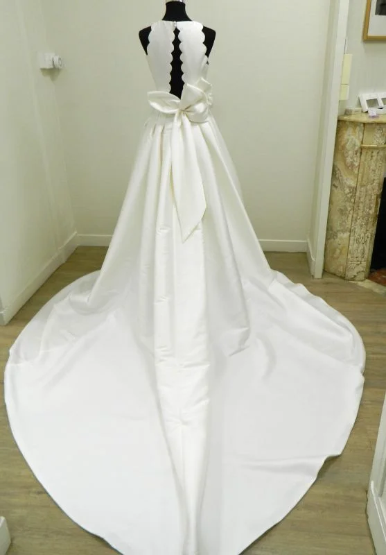 Robe de mariée princesse Evana de Rosa Clará vue de dos sur mannequin, dévoilant une ouverture dos nu verticale aux bords festonnés, un grand nœud en mikado et une immense traîne fluide.