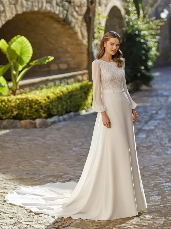Robe de mariée trapèze en crêpe Rosa Clara modèle Urim, vue de face avec une longue traîne sur une allée pavée. La robe présente un bustier en dentelle, une ceinture avec un bouton central et des manches longues transparentes.